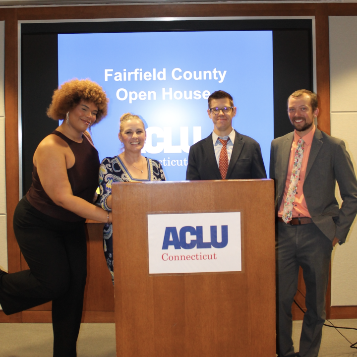 Behind a wooden podium with the ACLU-CT's logo, stands a Black-Taino women with a light brown afro on the left, a white woman with blonde hair to her right, a white man with dark hair and glasses to her right, and a white man with brown hair on the right.