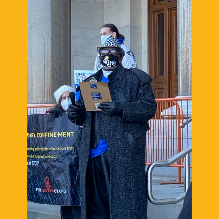 ACLUCT smart justice leader Curtis B. Hudson stands in front of the CT Capitol steps. He is wearing a black and white fedora, sunglasses, black mask, black trenchcoat, and blue Smart Justice shirt, and black gloves, and he is holding a clipboard.