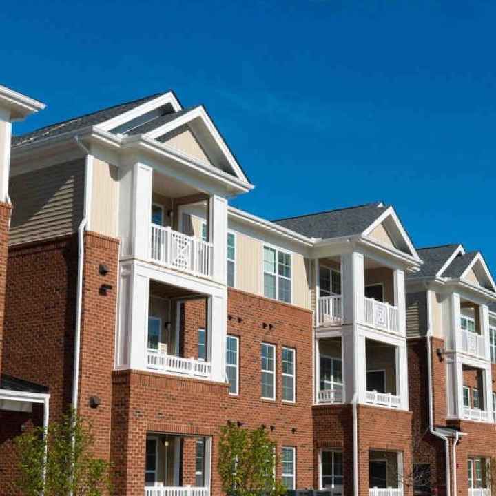 A block of apartments, white on top with balconies and brick below, stand in front of a blue sky.