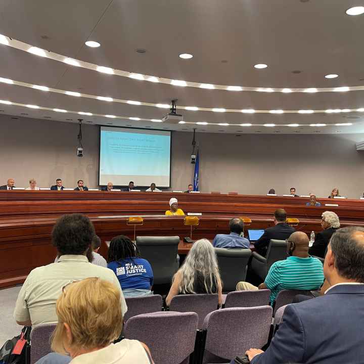 This photo captures a legislative forum about the fake traffic ticket scandal from the back of the room. Several people's backs are towards the camera with legislators in the background of the photo.