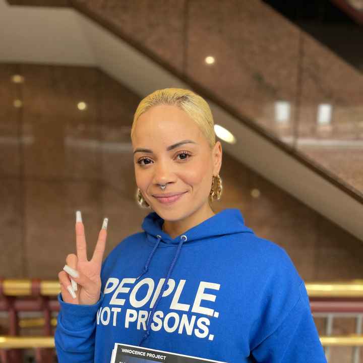 ACLU of CT Smart Justice leader Marquita Reale stands, smiling at the camera and holding up a peace sign. she is wearing a blue people not prisons hoodie