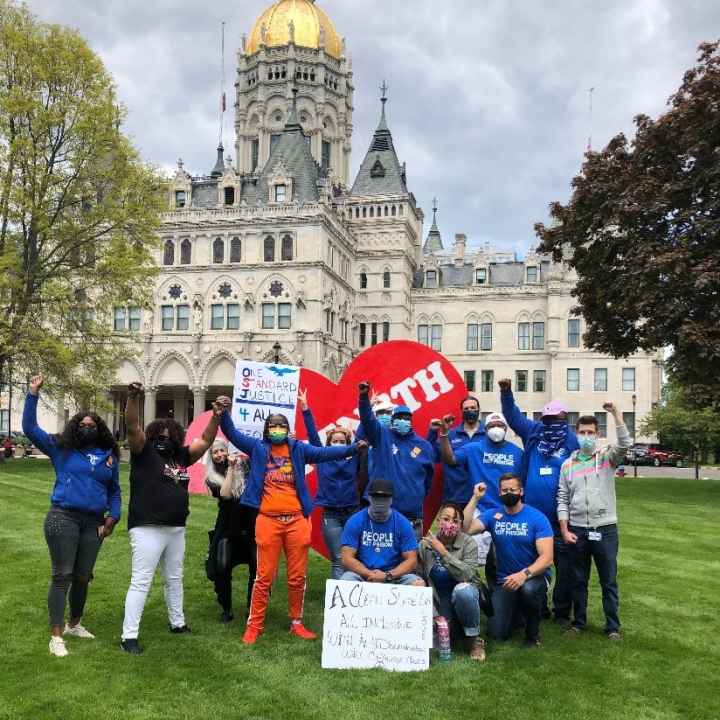 ACLUCT Smart Justice leaders, in blue people not prisons shirts, crouch and stand with a massive red heart that says "strength" in white letters. Behind them is the CT capitol building. They are masked and raising their fists in celebration and power.