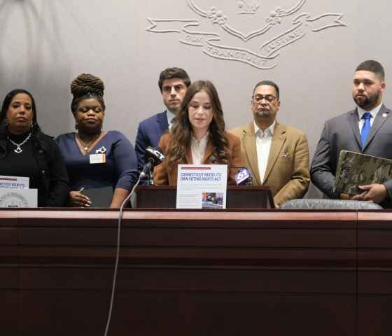 Jess Zaccagnino, a woman with long brown hair, stands behind a podium. In front of her is a sign that says "CT voting rights act." Behind her is a group of people, watching as she speaks