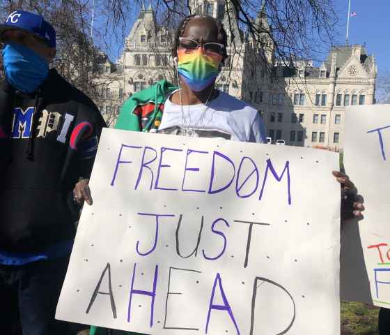 ACLU-CT Smart Justice leader Terri Ricks stands in front of the CT State Capitol Building. She is wearing sunglasses and a rainbow face mask, and she holds a white sign that says, in blue letters, "FREEDOM JUST AHEAD."