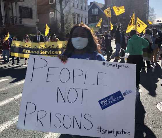 Smart Justice leader Shelby Henderson holds a white sign that says "PEOPLE NOT PRISONS" in blue ink. Behind her is a crowd waving yellow flags and carrying a banner. They are on a street in Hartford, marching.